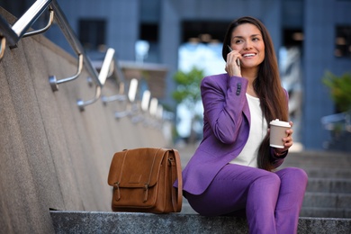 Beautiful businesswoman with cup of coffee talking on phone outdoors