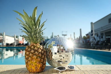 Shiny disco ball, pineapple and sunglasses on edge of swimming pool. Party items