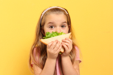 Photo of Cute little girl eating tasty sandwich on yellow background