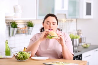Overweight woman eating sandwich instead of salad at table in kitchen. Healthy diet