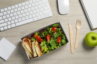 Photo of Container of tasty food, keyboard, apple, cutlery and notebook on white wooden table, flat lay. Business lunch