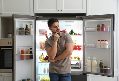 Thoughtful young man near open refrigerator in kitchen