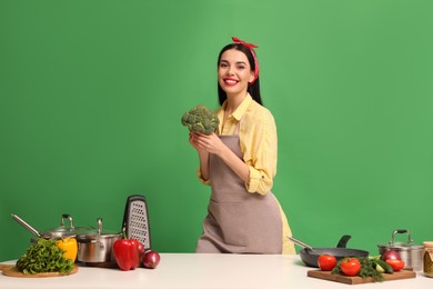 Young housewife with vegetables and different utensils on green background