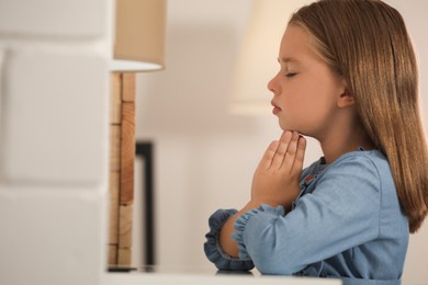 Cute little girl with hands clasped together praying at home. Space for text
