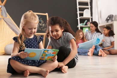Cute little children reading book on floor in kindergarten, space for text. Indoor activity
