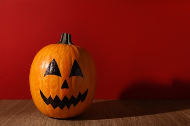 Pumpkin with drawn spooky face on wooden table against red background, space for text. Halloween celebration