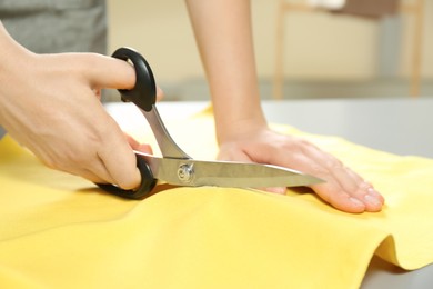 Photo of Seamstress cutting yellow fabric with scissors at workplace, closeup