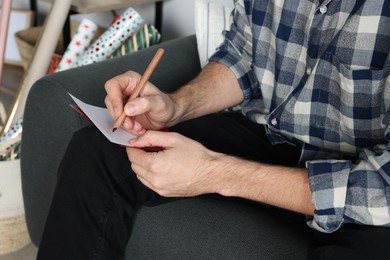 Man writing message in greeting card on sofa in room, closeup