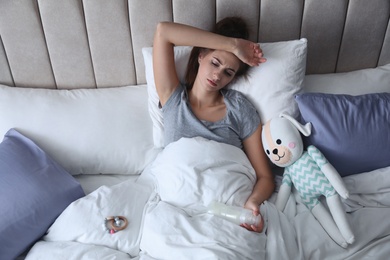 Photo of Exhausted young mother with toy and bottle of milk in bed at home, above view