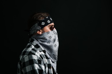 Fashionable young man with bandana covering his face on black background, space for text