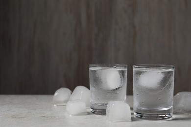 Shot glasses of vodka with ice cubes on light grey table against wooden background. Space for text