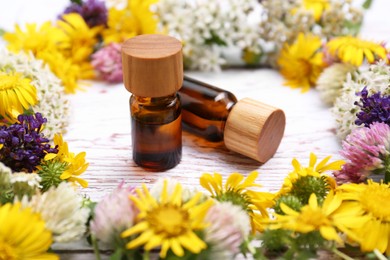 Bottles of essential oils surrounded by beautiful flowers on white wooden table, closeup