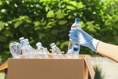 Woman putting used plastic bottle into cardboard box outdoors, closeup. Recycle concept
