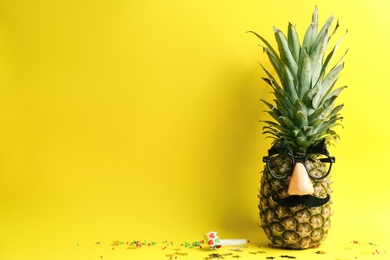 Photo of Pineapple with mask, party horn and confetti on yellow background, space for text. Creative concept