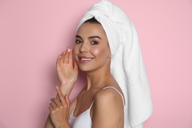 Beautiful young woman with towel applying cream on face against pink background