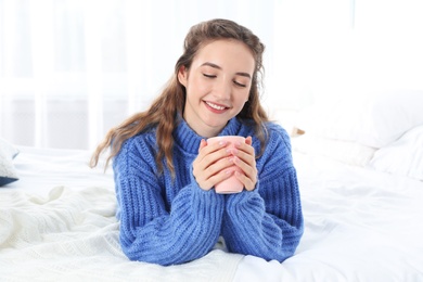Beautiful teenage girl in warm cozy sweater with cup of hot drink on bed at home