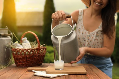 Smiling woman pouring fresh milk from can into glass at wooden table outdoors, closeup