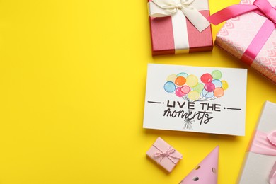Photo of Life-affirming phrase Live The Moments. Flat lay composition with card and gift boxes on yellow background, space for text