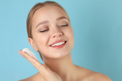 Young woman washing face with cleansing foam on light blue background. Cosmetic product