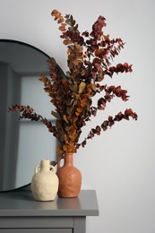 Vases, dried eucalyptus branches and stylish round mirror on grey table near white wall indoors. Interior design
