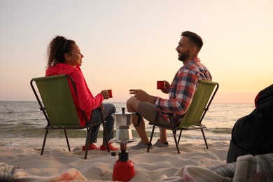 Couple resting in camping chairs near sea at sunset
