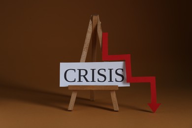 Wooden easel with word Crisis and descending red arrow on brown background