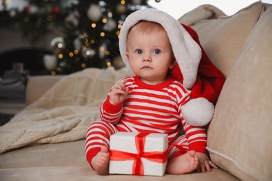 Cute baby in Santa hat with Christmas gift on sofa at home