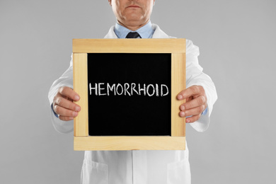 Doctor holding blackboard with word HEMORRHOID on light grey background, closeup