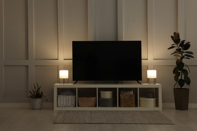 Photo of Modern TV on cabinet, lamps and beautiful houseplants near white wall in room. Interior design