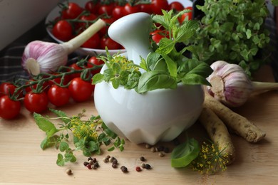 Photo of Mortar with fresh herbs near garlic, horseradish roots, black peppercorns and cherry tomatoes on wooden table, closeup