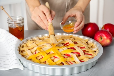 Woman applying liquid egg onto traditional English apple pie with brush at light grey table, closeup