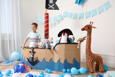 Photo of Cute little boy playing with pirate cardboard ship and toys at home. Child's room interior