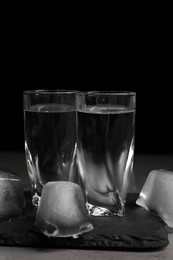 Shot glasses with vodka and ice cubes on grey tale against black background, closeup