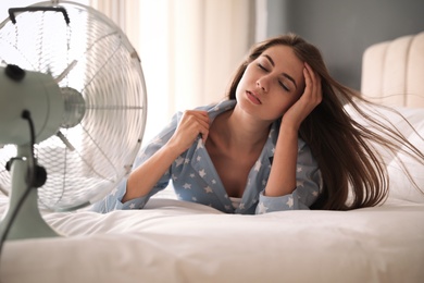 Woman enjoying air flow from fan on bed in room. Summer heat