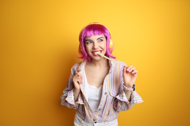 Fashionable young woman in colorful wig with headphones chewing bubblegum on yellow background