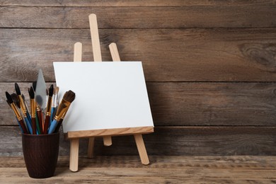 Easel with blank canvas and brushes on wooden table. Space for text