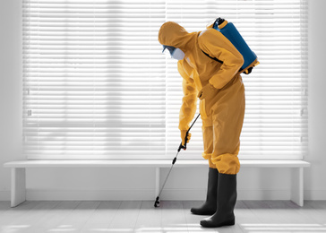 Male worker in protective suit spraying insecticide on wooden floor indoors, space for text. Pest control