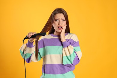 Stressed young woman with flattening iron on yellow background. Hair damage