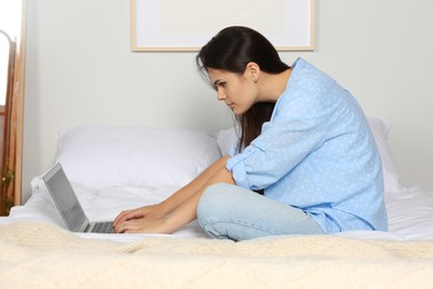 Photo of Young woman with bad posture using laptop while sitting on bed at home. Symptom of scoliosis