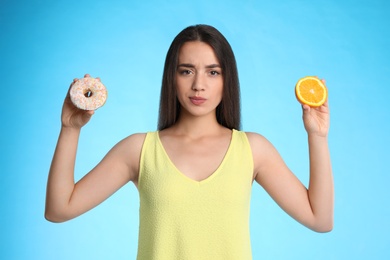 Doubtful woman choosing between orange and doughnut on light blue background