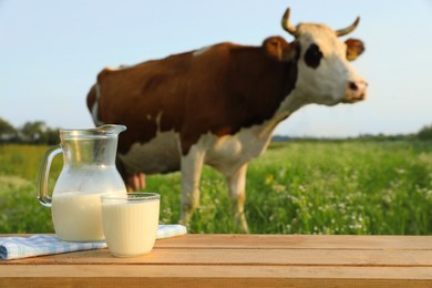 Glass with jug of milk on wooden table and cow grazing in meadow