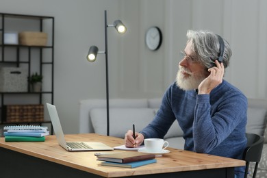 Middle aged man with laptop, notebook and headphones learning at table indoors