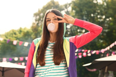 Photo of Beautiful young woman blowing chewing gum and showing peace gesture outdoors