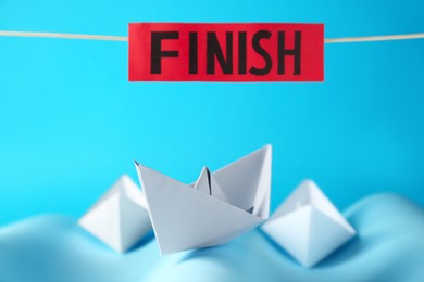 Photo of Competition concept. Paper boats among waves and red sign with word Finish on light blue background