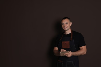 Portrait of happy young waiter with notebook on brown background, space for text