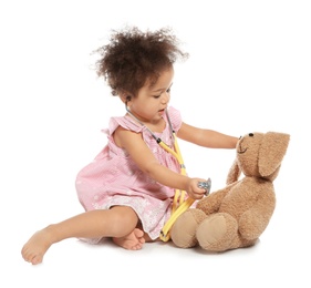 Cute African American child imagining herself as doctor while playing with stethoscope and toy bunny on white background