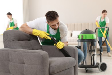 Professional janitor in uniform vacuuming armchair indoors