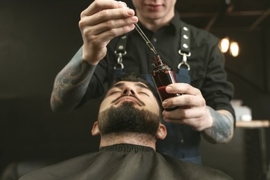 Image of Hairdresser with beard oil near client in barbershop, closeup. Professional shaving service