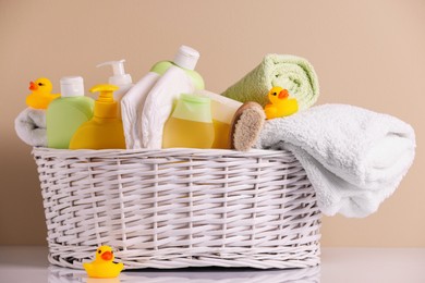 Wicker basket with baby cosmetic products, bath accessories and rubber ducks on white table against beige background