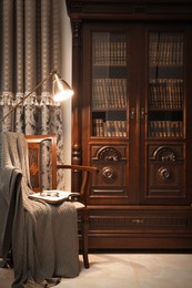 Comfortable armchair with book, blanket and lamp near wooden bookcase in library